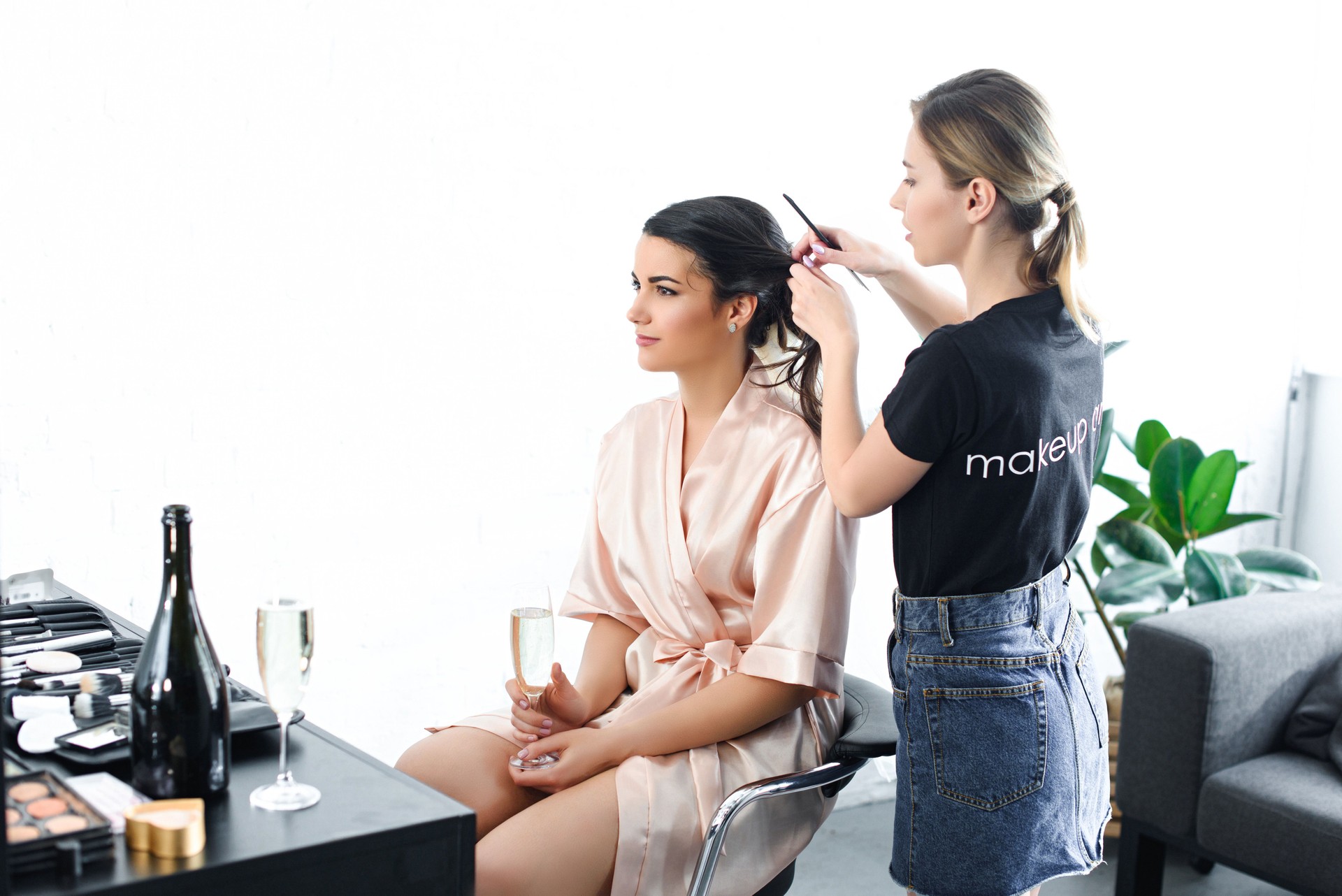 young hairstylist doing hairstyle for bride in silk bathrobe with glass of champagne